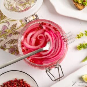 A glass jar filled with pickled red onions and pink peppercorns sits open on a white surface. A fork rests inside the jar. A floral napkin, a white plate with food, and a small bowl of peppercorns are nearby.