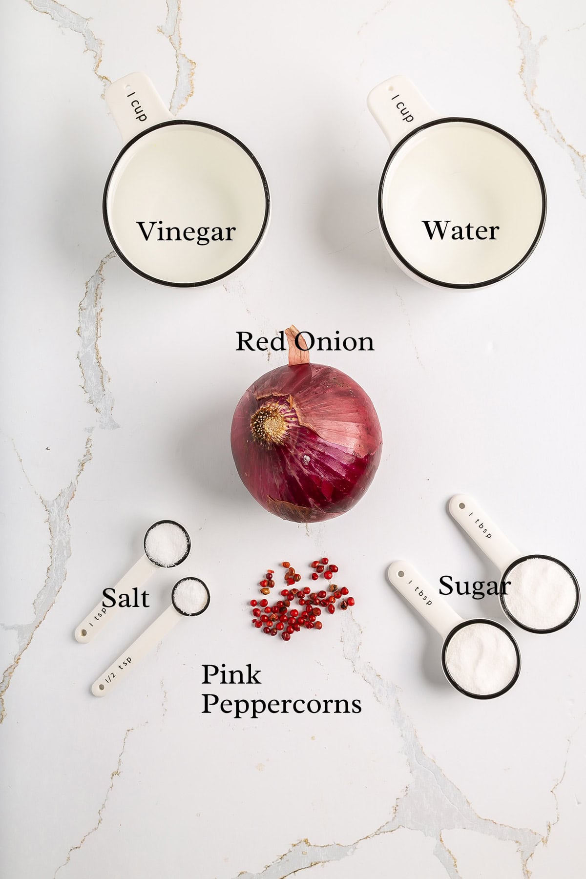 Overhead view of ingredients on a white surface: two measuring cups labeled vinegar and water, a whole red onion, measuring spoons with salt and sugar, and a small pile of pink peppercorns.