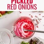 A glass jar filled with sliced pickled red onions sits open on a white table next to a fork and plates of salad greens. The text Pickled Red Onions appears above the jar, with a Lana’s Cooking logo in the corner.