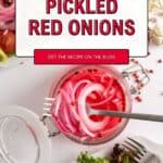 A glass jar filled with pickled red onions sits open on a white table, surrounded by plates of salad with lettuce and tomato. A fork rests in the jar. The text reads Pickled Red Onions and GET THE RECIPE ON THE BLOG.