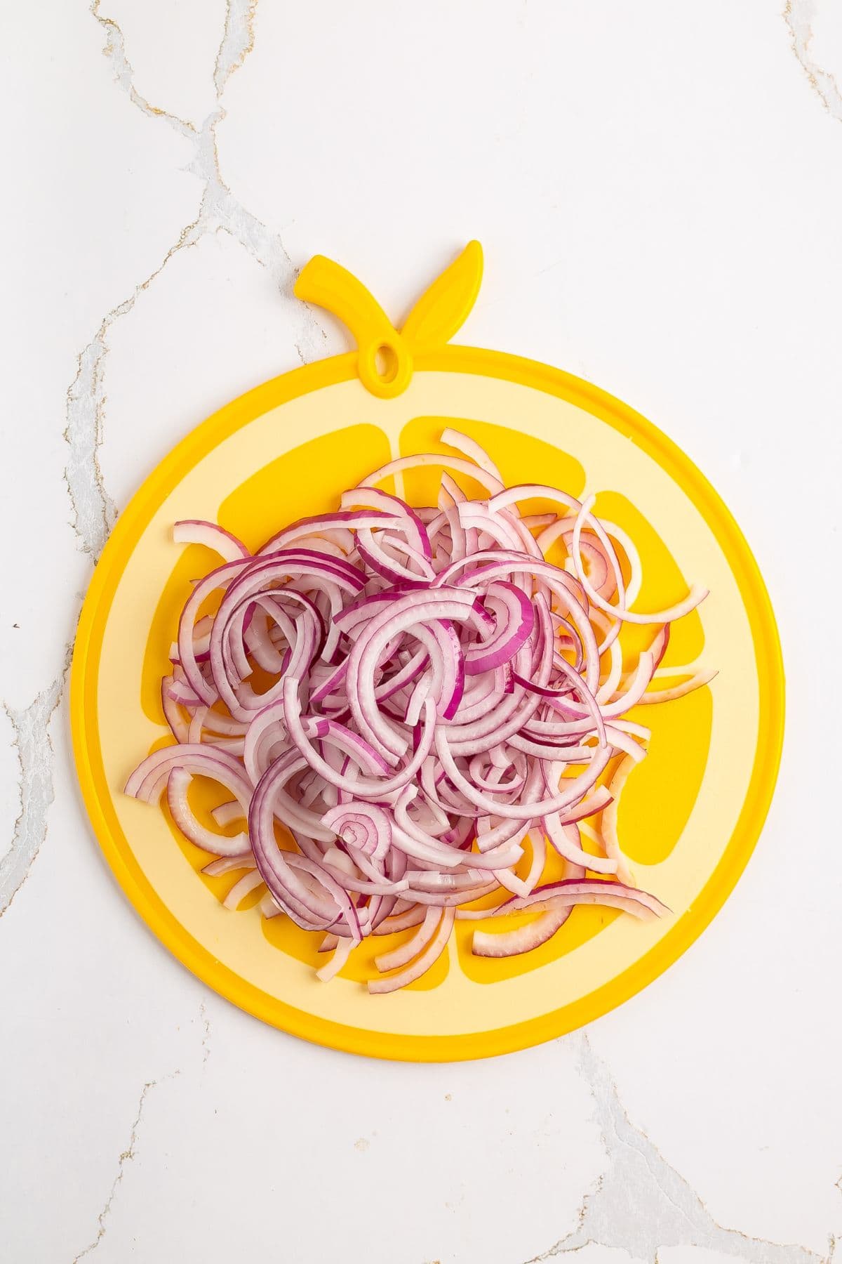 Thinly sliced red onions are arranged in a pile on a yellow cutting board shaped like a fruit slice, placed on a white surface with visible cracks.