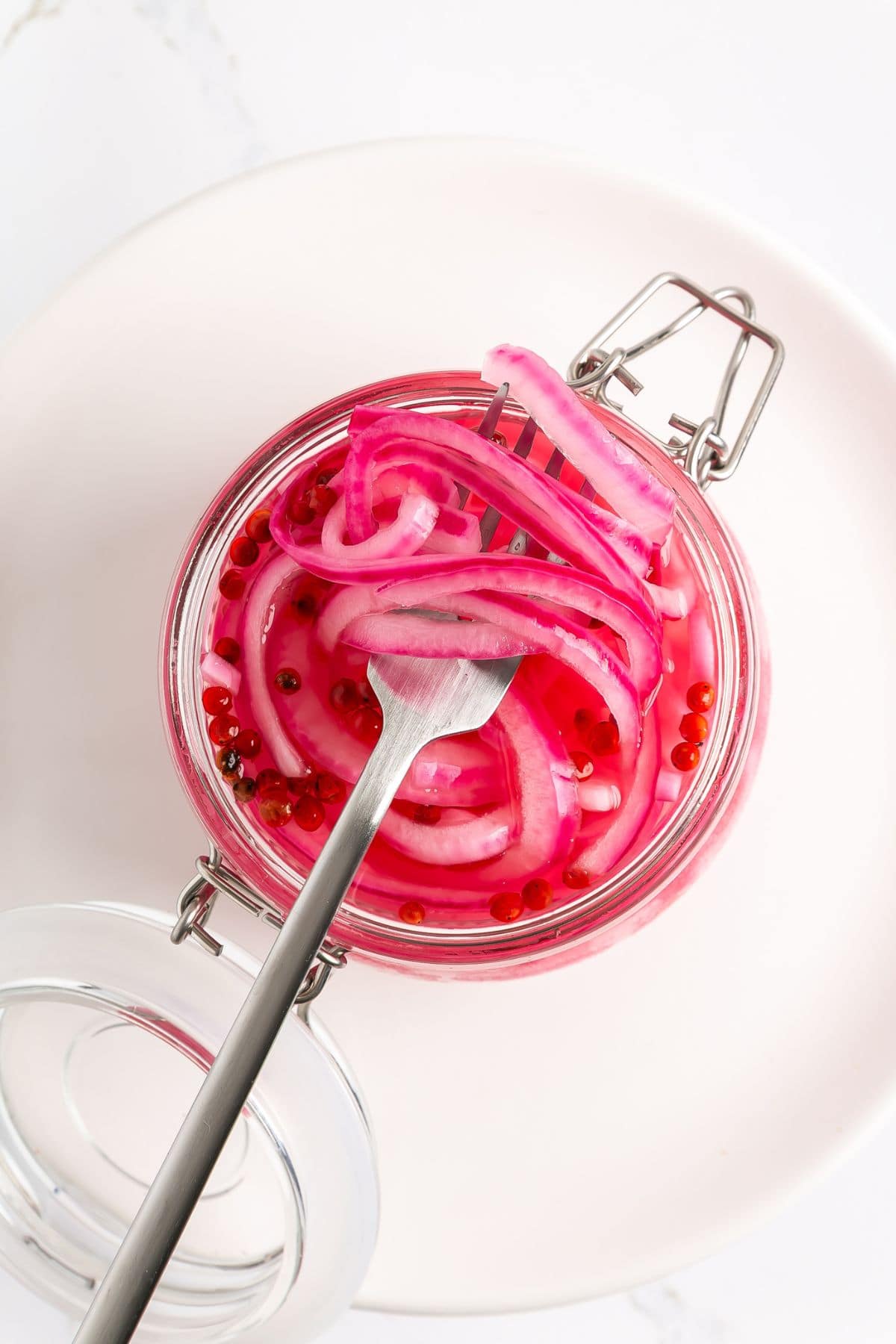 A jar of pickled red onions with pink peppercorns sits on a white plate, with a fork lifting some onions from the open jar.