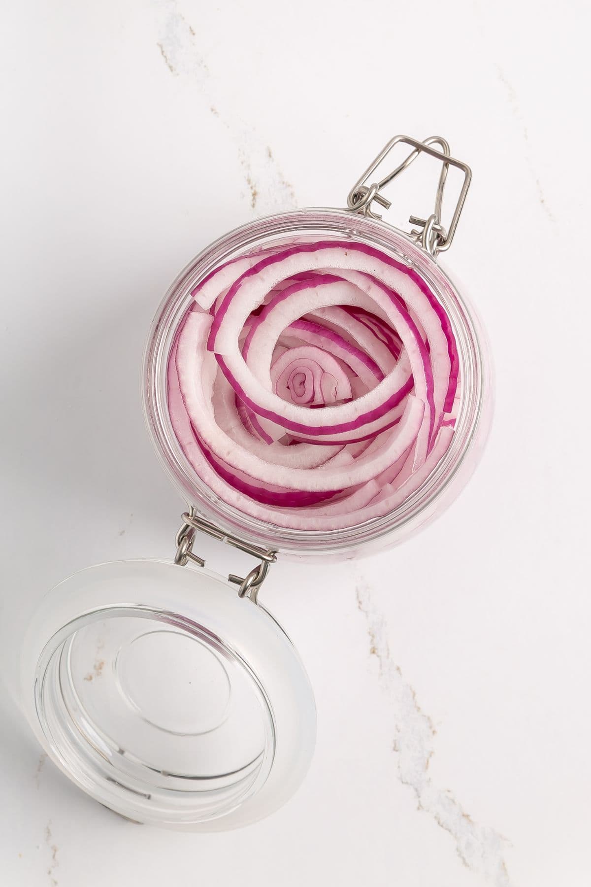 A glass jar with a latch lid contains thinly sliced red onions arranged in circular layers, viewed from above on a white marble surface.