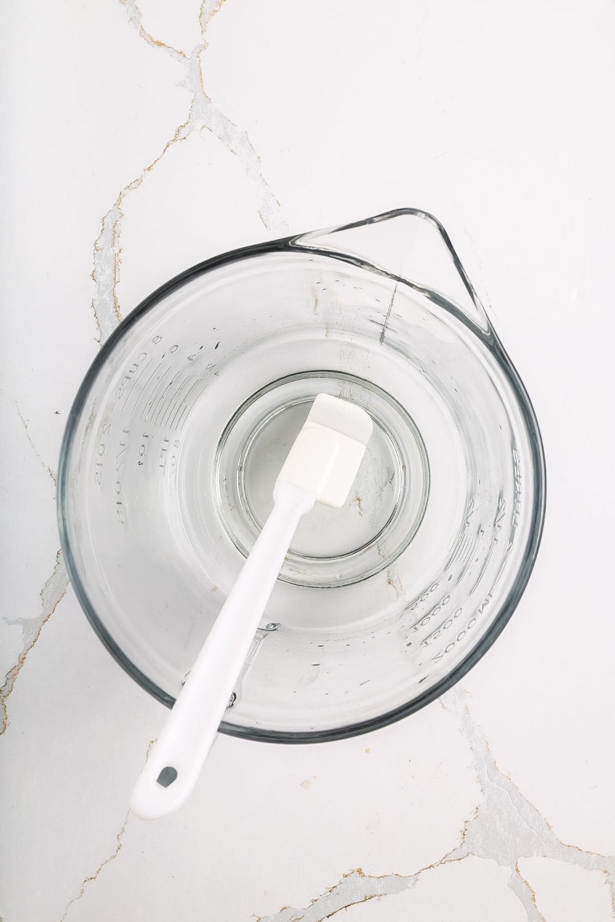 A clear glass measuring cup with a white silicone spatula inside, placed on a white surface with faint gray cracks.