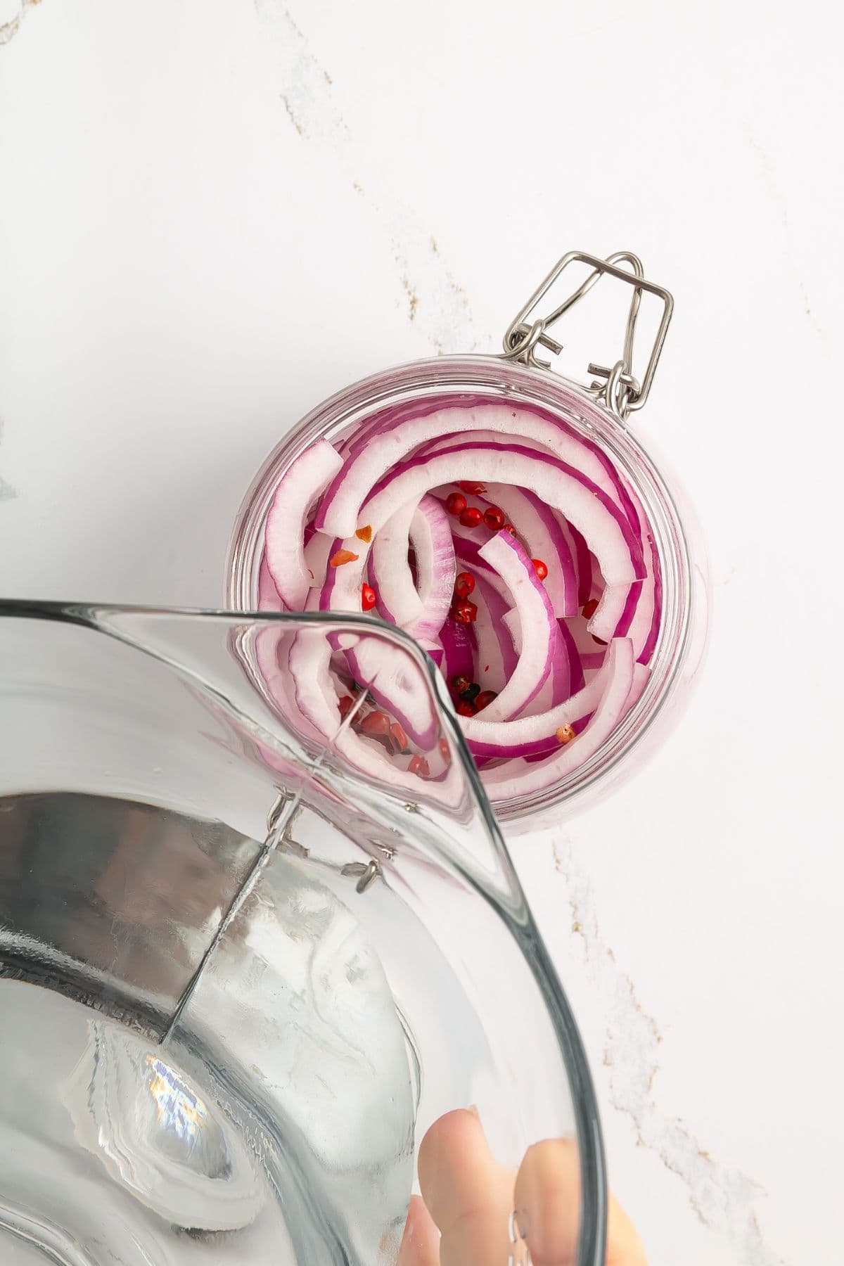 A hand pours clear liquid from a glass pitcher into a jar filled with sliced red onions and pink peppercorns, placed on a white marble surface.