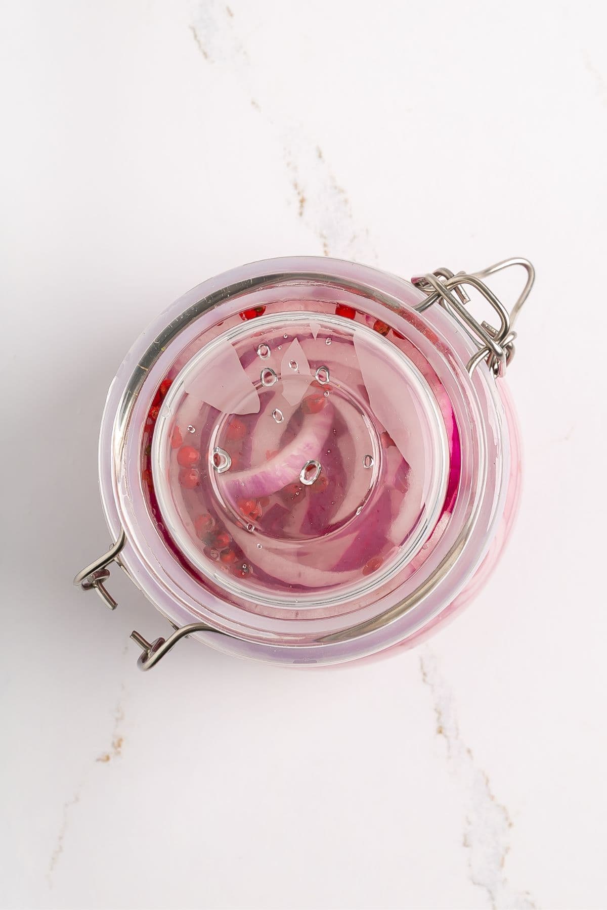 A glass jar with a metal clasp lid is filled with sliced red onions submerged in clear pickling liquid, viewed from above on a white marble surface.