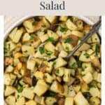 A bowl of roasted potato salad with garlic, rosemary, and parsley, with a serving spoon.