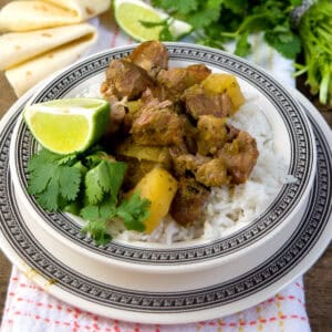 A plate of white rice topped with chunks of stewed pork and potatoes, garnished with a lime wedge and cilantro, sits on a decorative plate. Flour tortillas and fresh herbs are visible in the background.