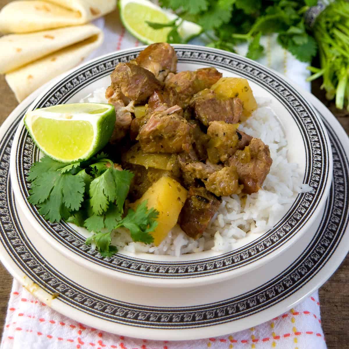 A plate of white rice topped with chunks of stewed pork and potatoes, garnished with a lime wedge and cilantro, sits on a decorative plate. Flour tortillas and fresh herbs are visible in the background.