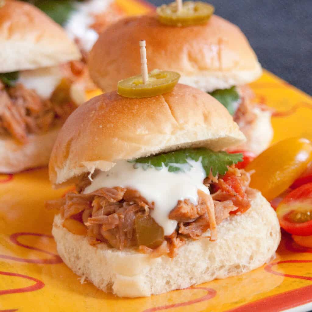 Two pulled pork sliders on soft buns are topped with white sauce, cilantro, and sliced jalapeño, secured with toothpicks. The sliders are on a yellow platter with sliced peppers on the side.