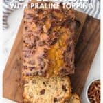 Sliced apple bread with pecan praline topping on a wooden board, next to a small bowl of pecans.