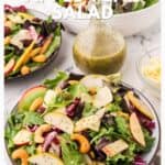 Salad with apple slices, cashews, greens, and a jar of lemon poppy seed dressing on a table.