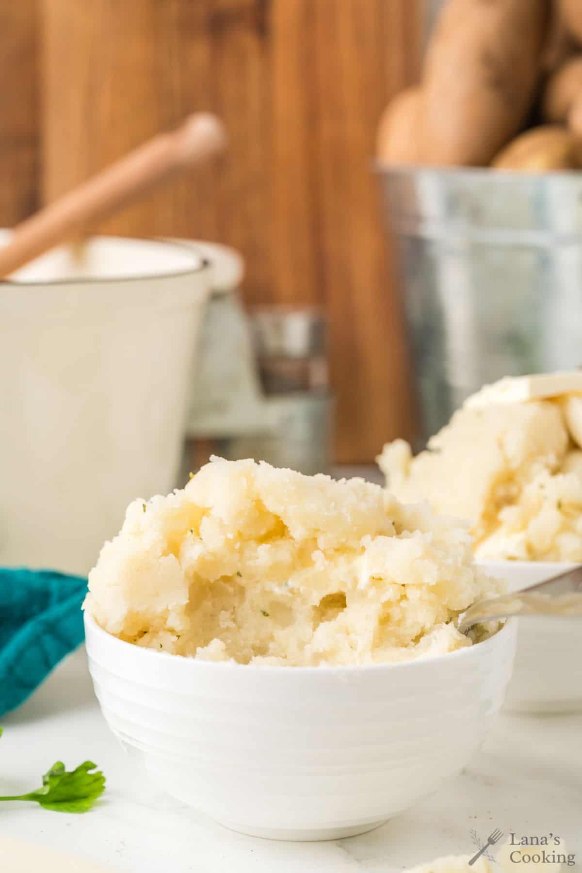 A white bowl filled with creamy mashed potatoes, with a spoon beside it on a white surface.