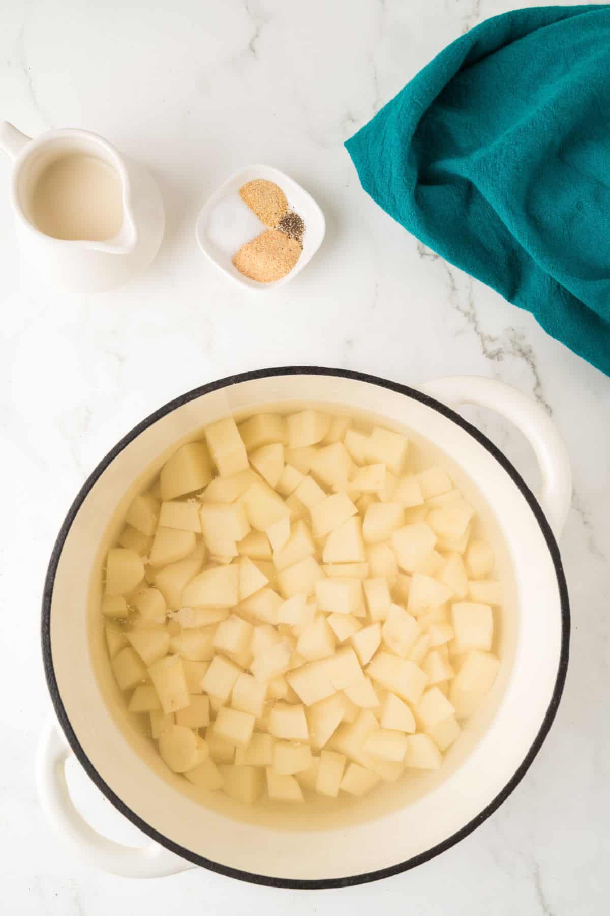 A pot of diced potatoes in water, with a small pitcher, spices, and a teal towel nearby on a marble counter.