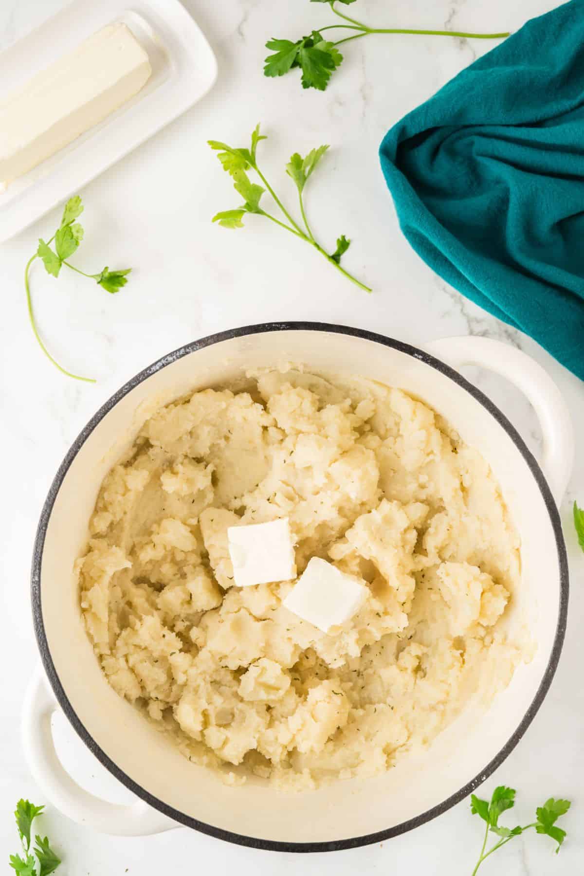 Pot of mashed potatoes with butter on top, surrounded by parsley and a teal cloth on a white surface.