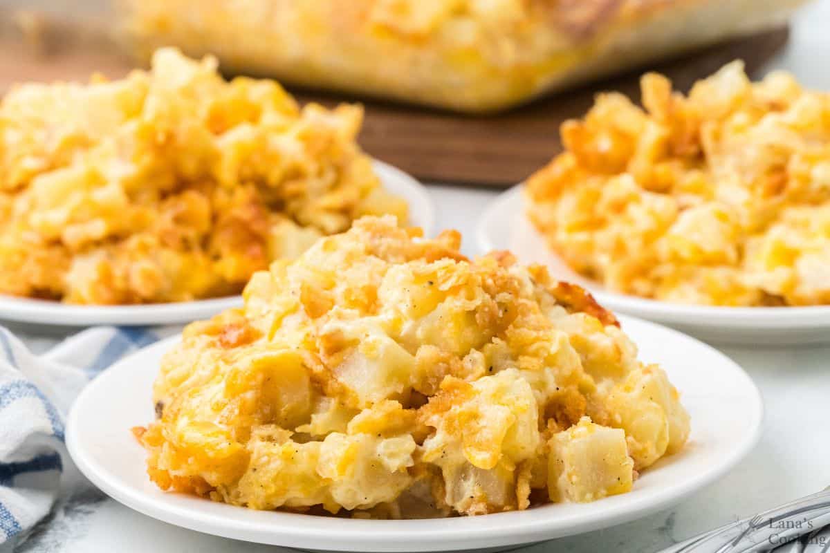 Three white plates with servings of cheesy hashbrown casserole, topped with a golden, crispy breadcrumb crust.