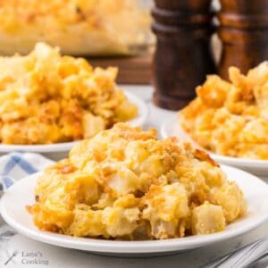 Close-up of cheesy hash brown casserole served on white plates, with a casserole dish and pepper mills in the background.
