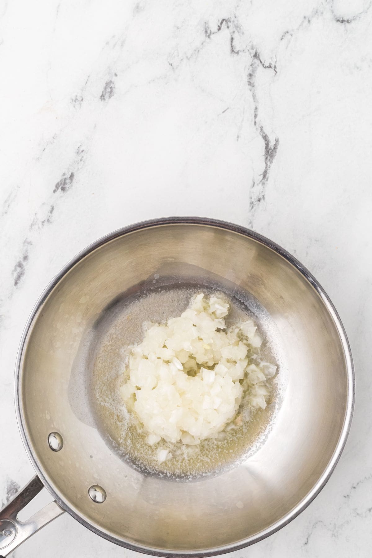 Chopped onions cooking in melted butter in a stainless steel saucepan on a white marble surface.