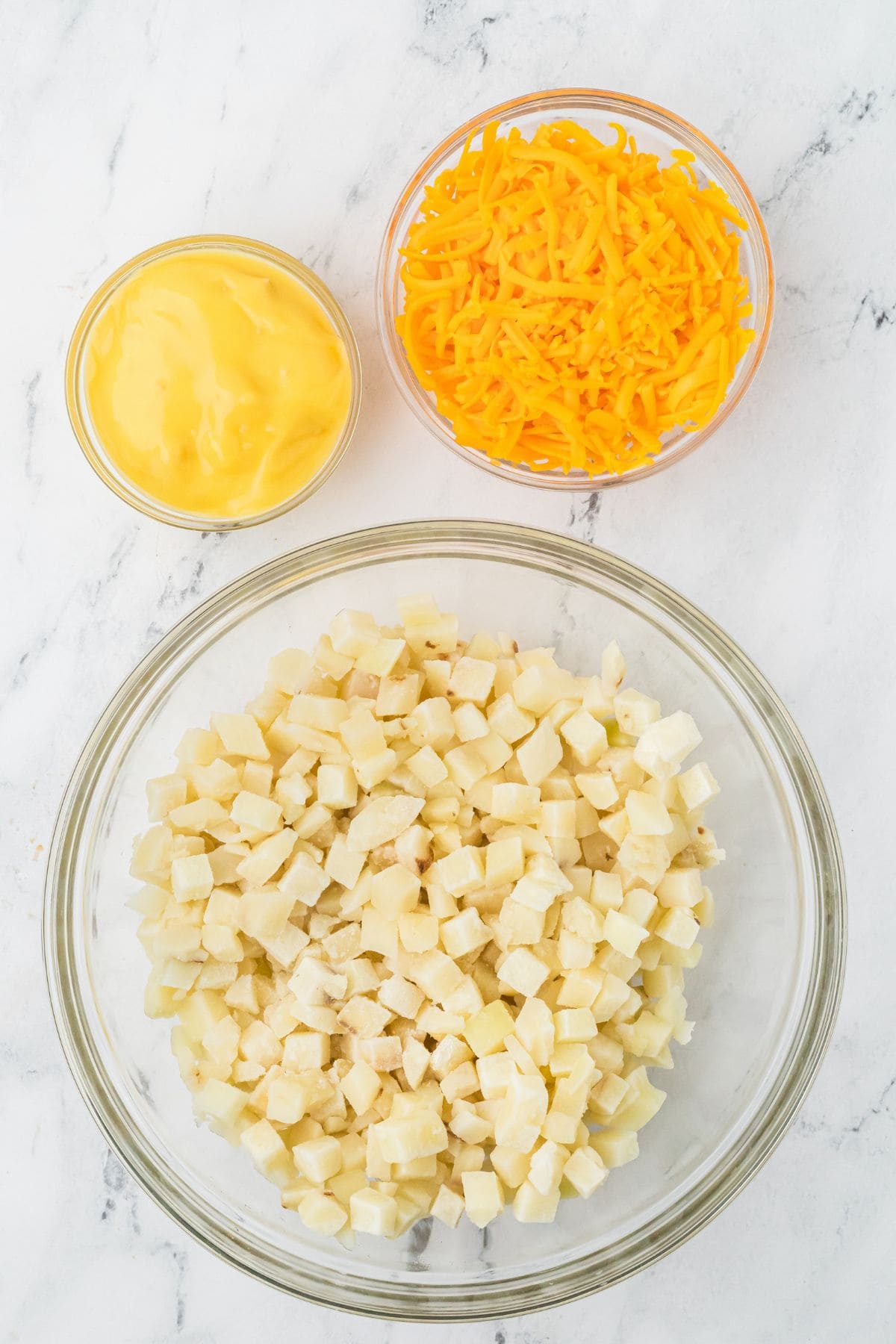 Bowls of diced potatoes, shredded cheddar cheese, and cheese sauce on a marble surface.