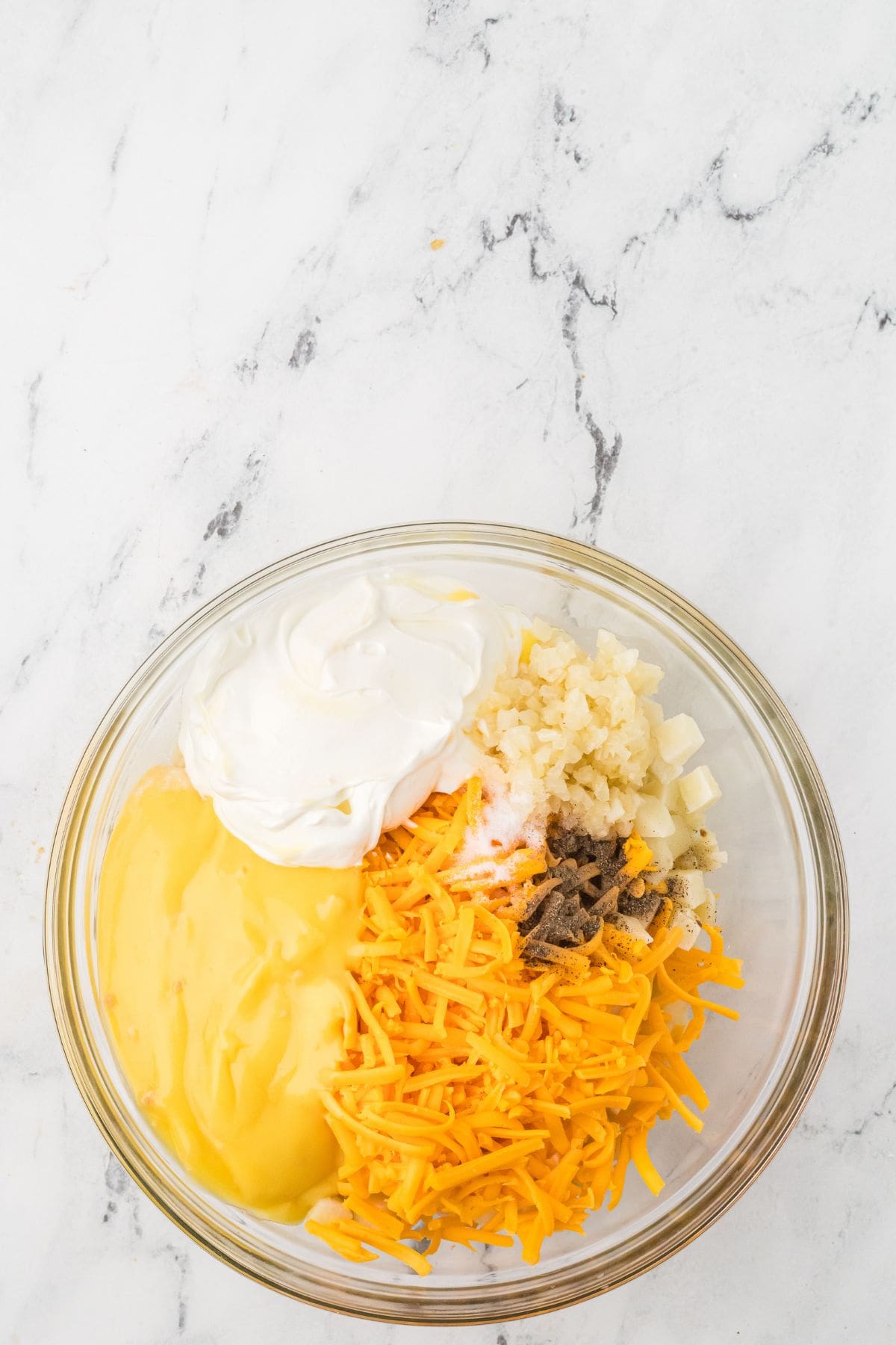 Glass bowl with shredded cheese, sour cream, chopped onions, condensed soup, and seasonings on a marble surface.