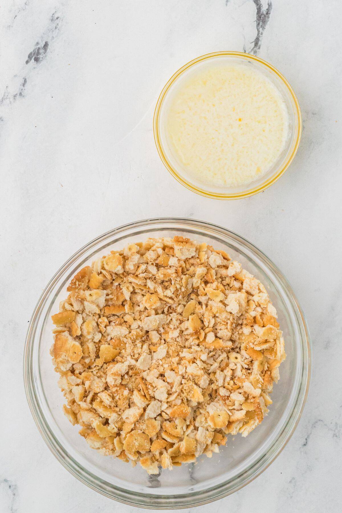 A bowl of crushed crackers and a bowl of melted butter on a white marble surface.
