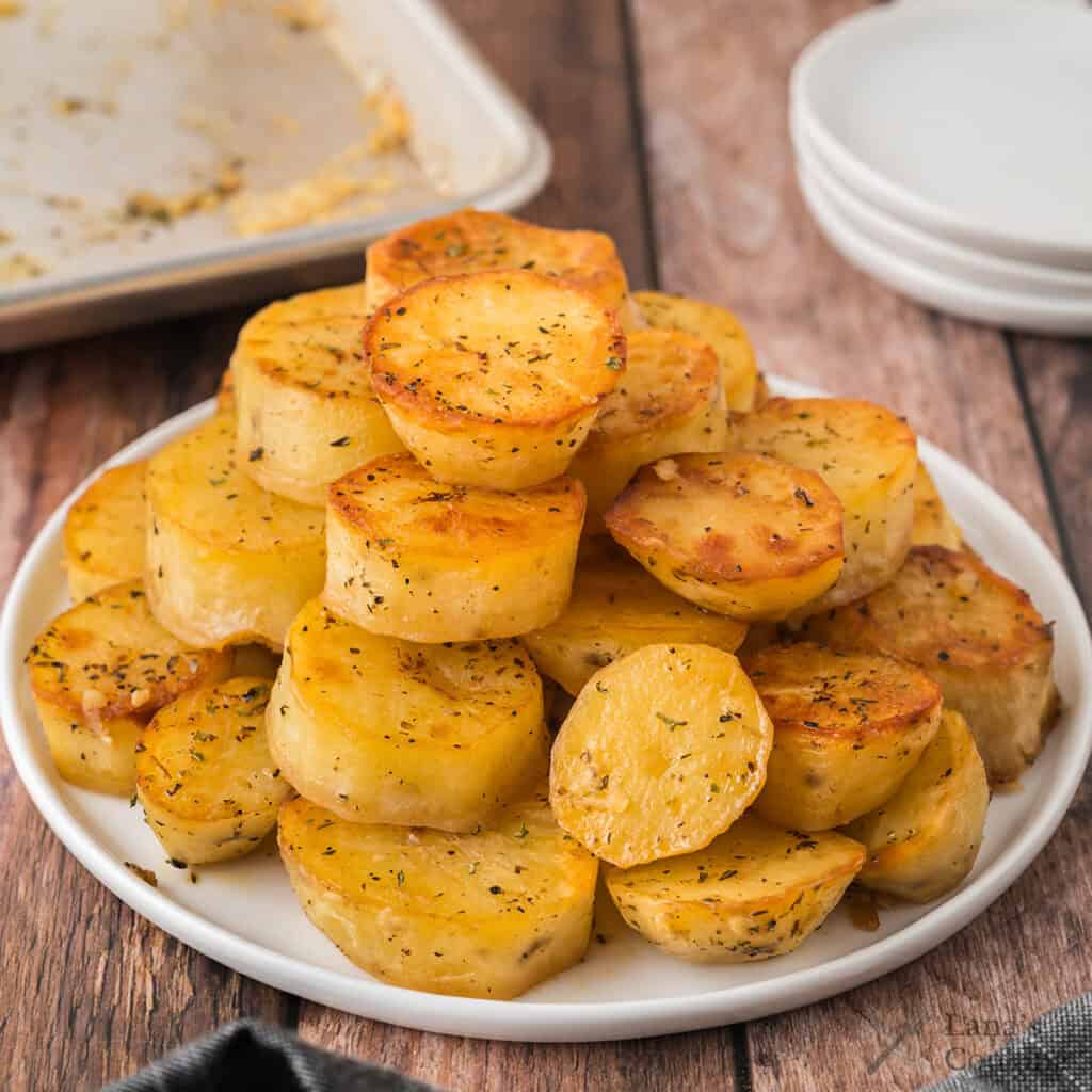 A white plate stacked with golden roasted potatoes, seasoned with herbs, on a wooden table.