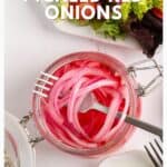 A jar of pickled red onions with a fork, surrounded by plates of fresh greens and a small dish.