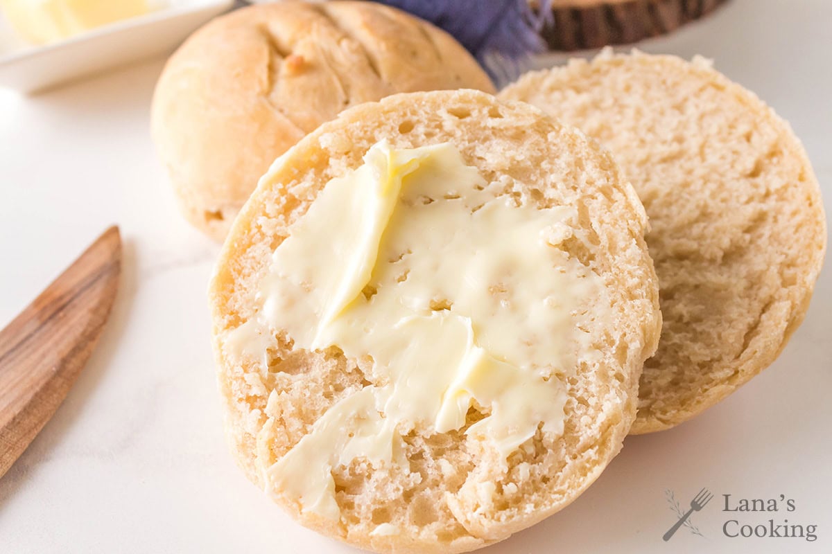 A split roll with butter spread on one half sits on a white surface, with another biscuit and a butter dish partially visible in the background. The text Lanas Cooking appears in the bottom right corner.