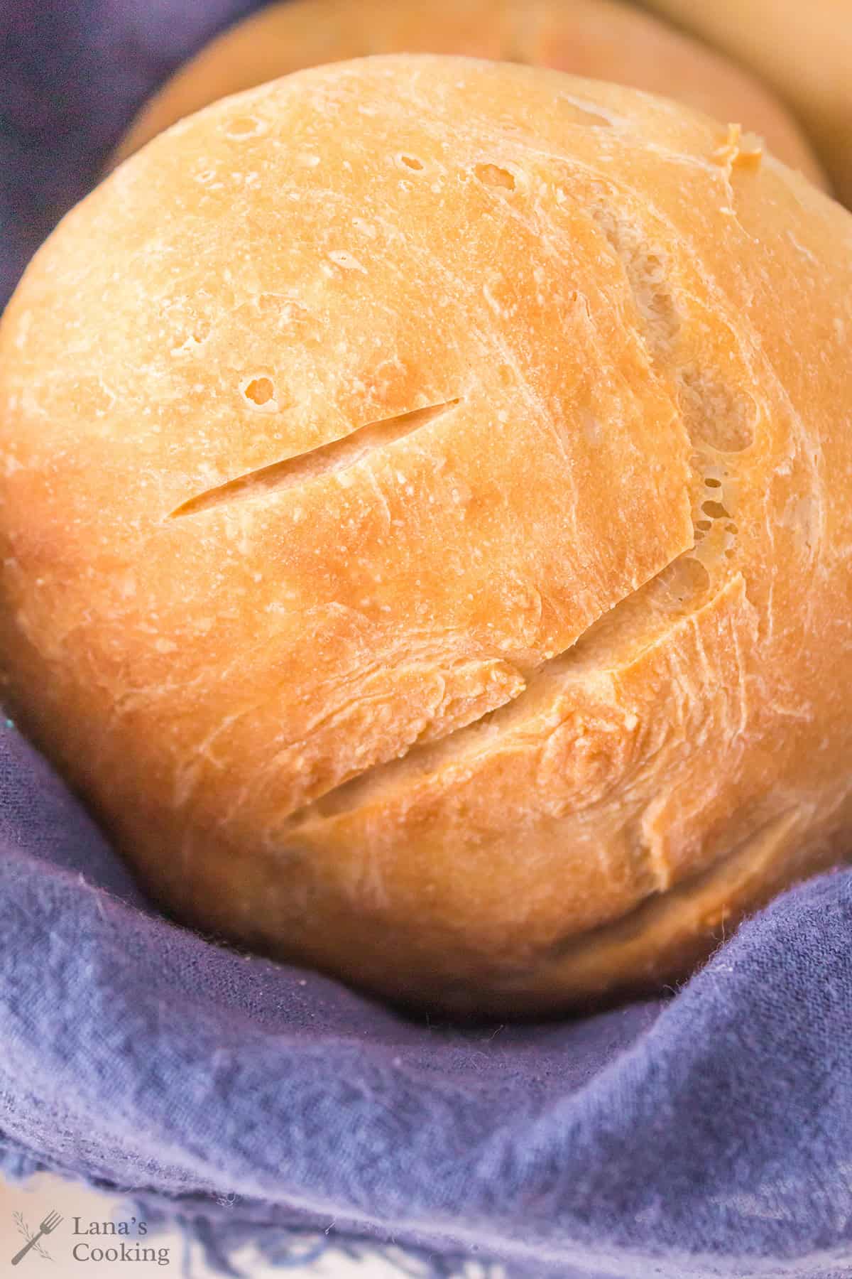 A golden-brown roll with two slashes on the crust rests on a dark blue cloth. The bread has a slightly crispy texture and a rustic appearance.