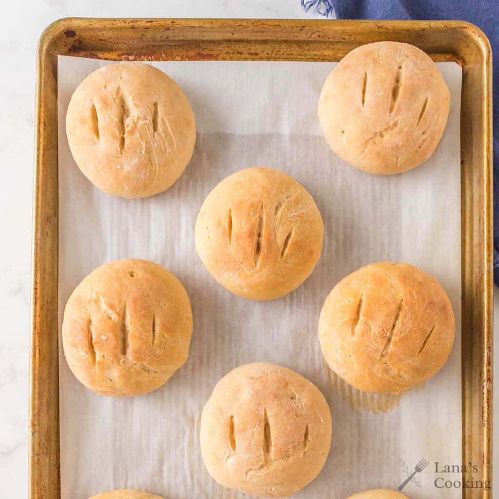 A baking tray lined with parchment paper holds seven round, golden-brown bread rolls, each with three shallow slashes on top. The tray is placed on a white surface. “Lana’s Cooking” logo is visible in the lower right corner.