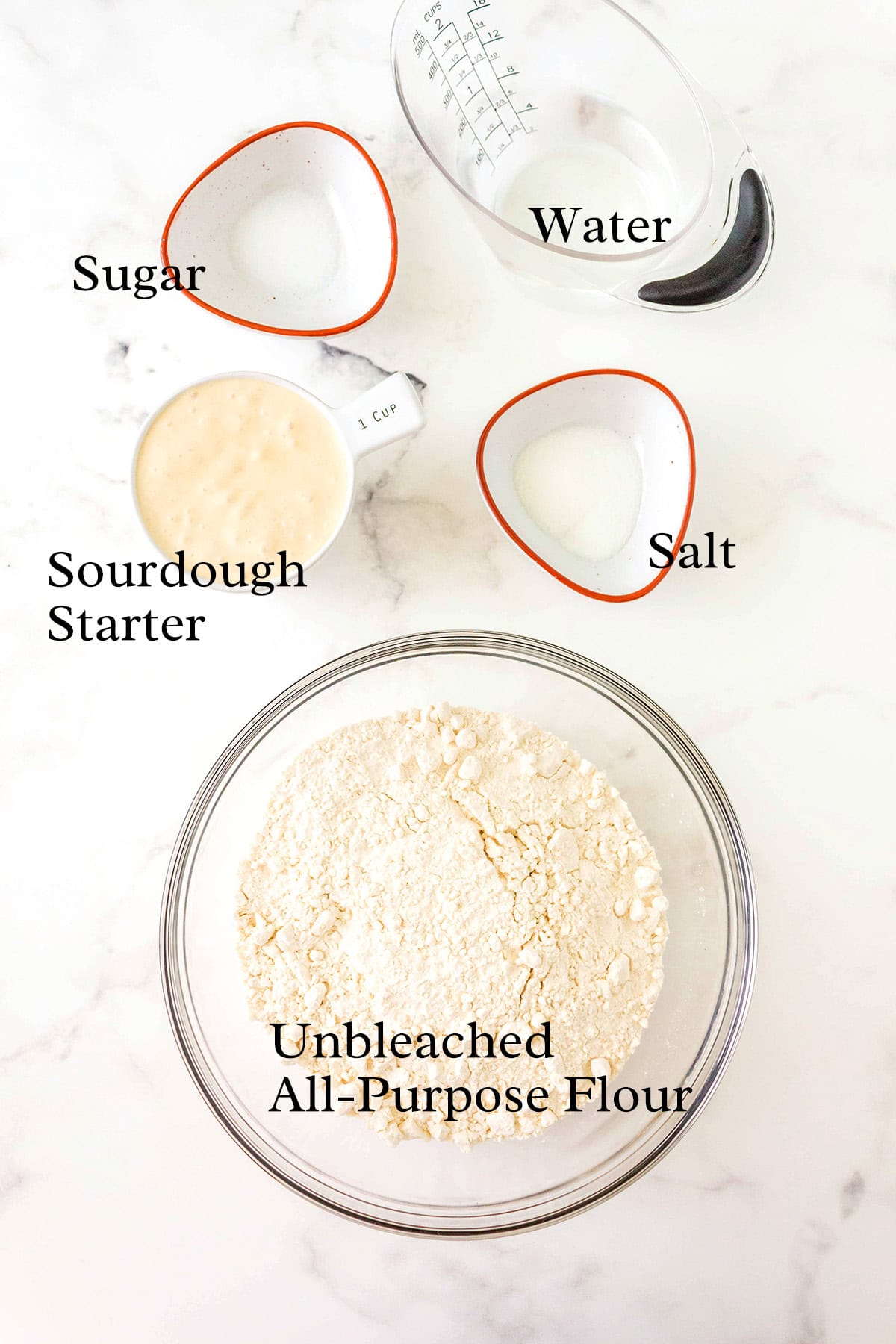 A glass bowl of unbleached all-purpose flour is on a white surface, surrounded by bowls containing sourdough starter, sugar, salt, and a measuring cup of water. Each ingredient is labeled.