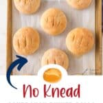 A baking sheet with six round, golden-brown dinner rolls on parchment paper. Text below reads No Knead Sourdough Dinner Rolls — Get the Recipe — lanascooking.com. An arrow points to the rolls.