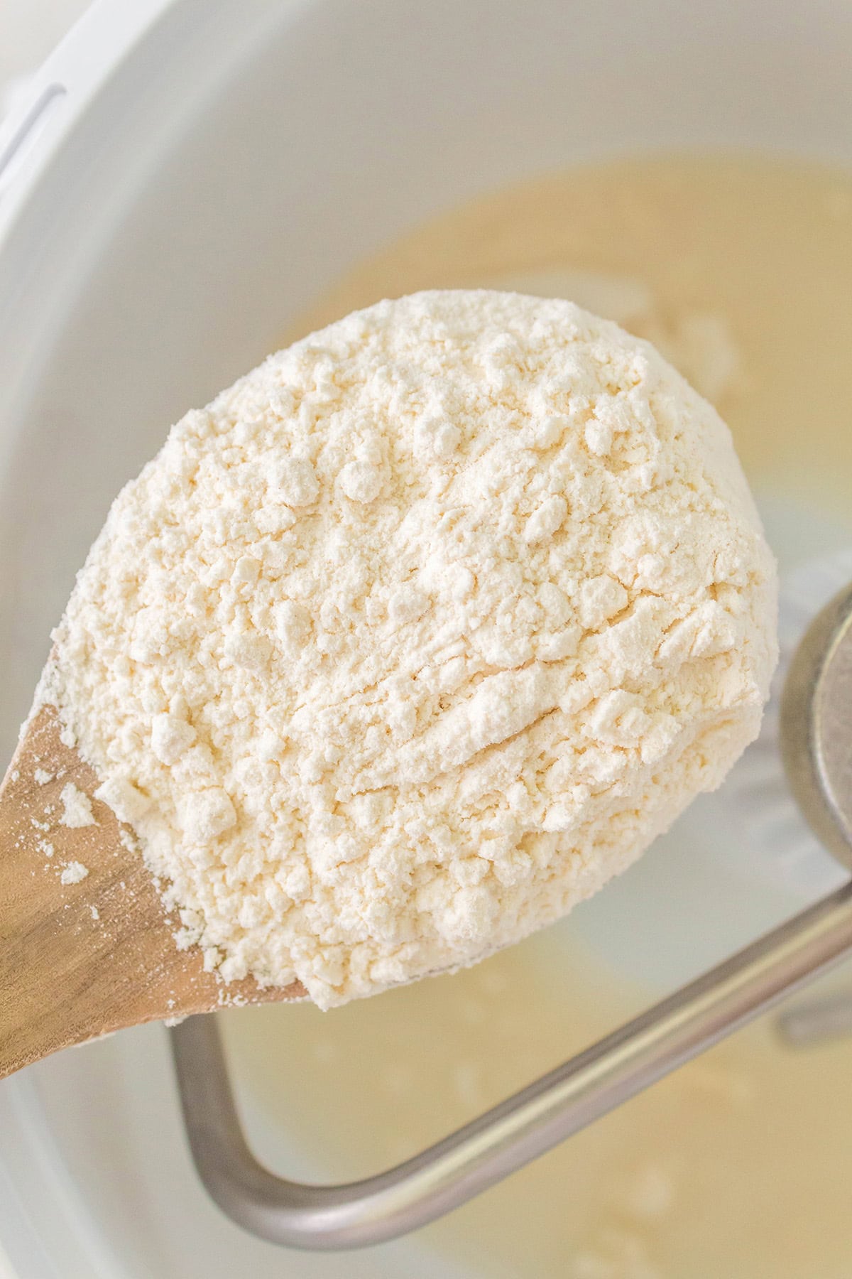 A wooden spoon holding a mound of flour over a mixing bowl with a metal dough hook attachment visible inside the bowl.