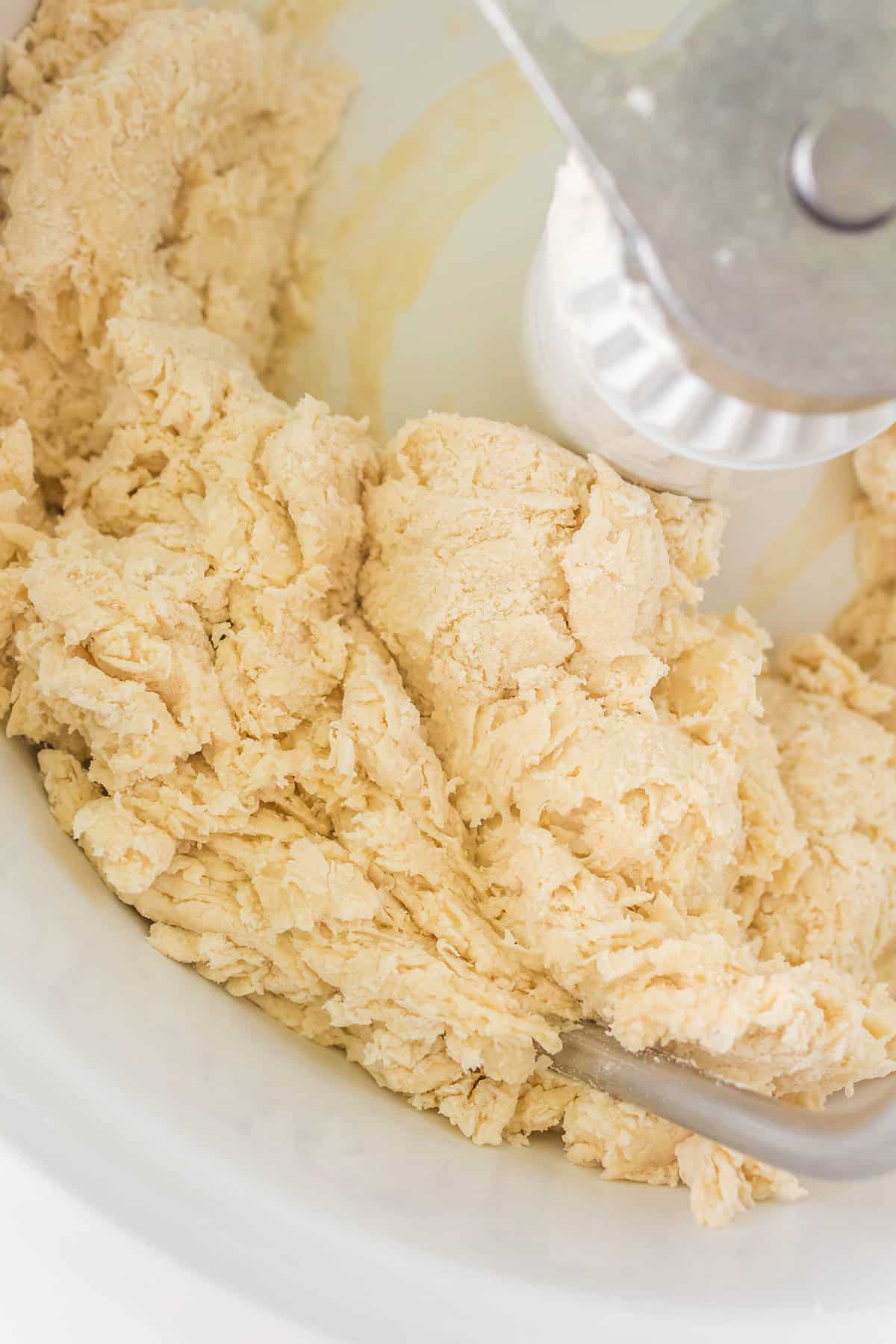 A close-up view of raw dough being mixed in a white stand mixer bowl with a metal attachment visible.