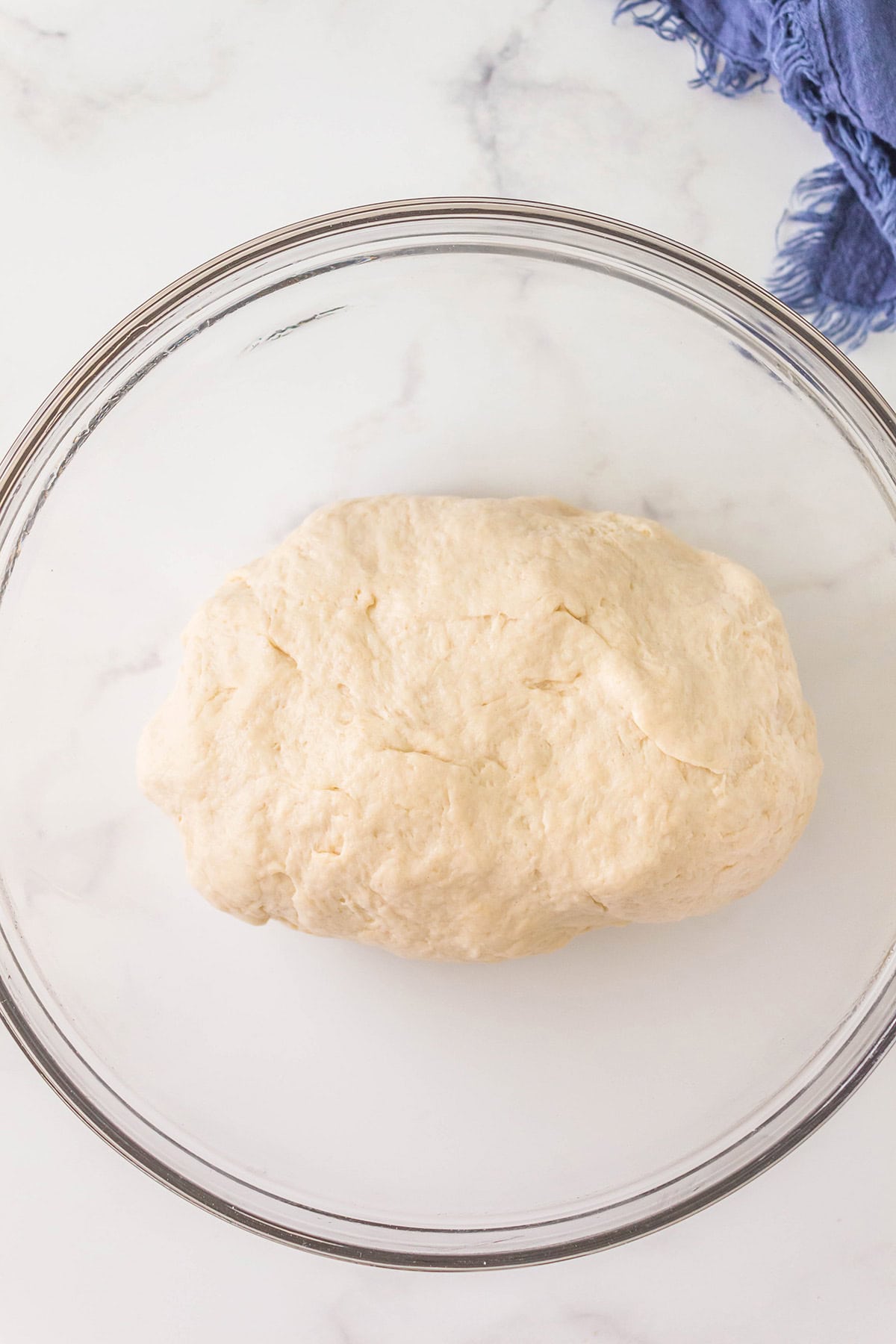 A ball of raw dough sits in a clear glass bowl on a white marble surface. A piece of blue cloth is partially visible in the upper right corner.