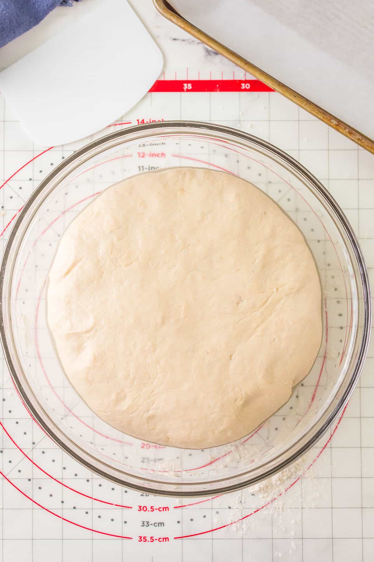A large, round ball of dough is rising in a clear glass bowl on a red and white measurement mat. A white dough scraper and a parchment-lined baking sheet are nearby.