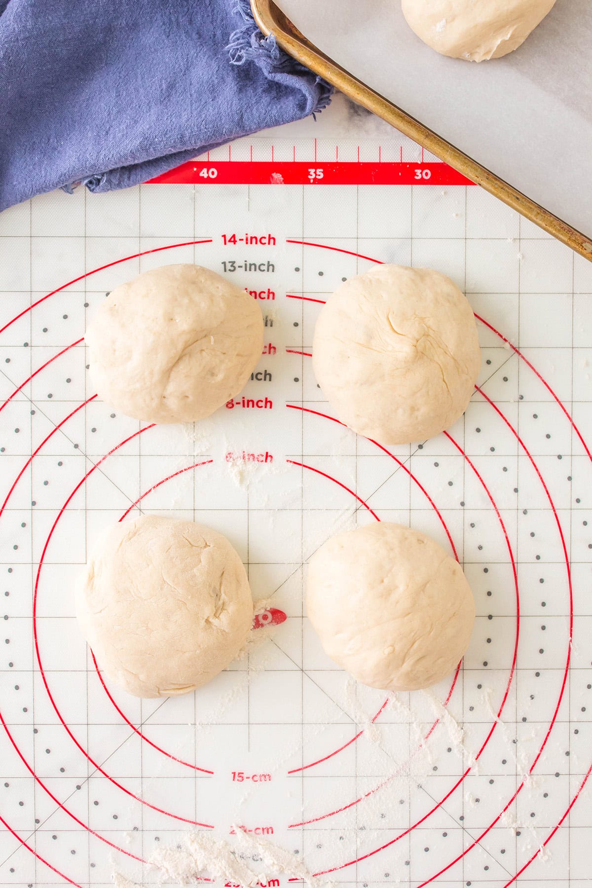 Four dough balls rest on a white silicone pastry mat with red concentric circles and measurement markings. A blue cloth is in the top left corner, and a baking sheet with more dough is in the top right.