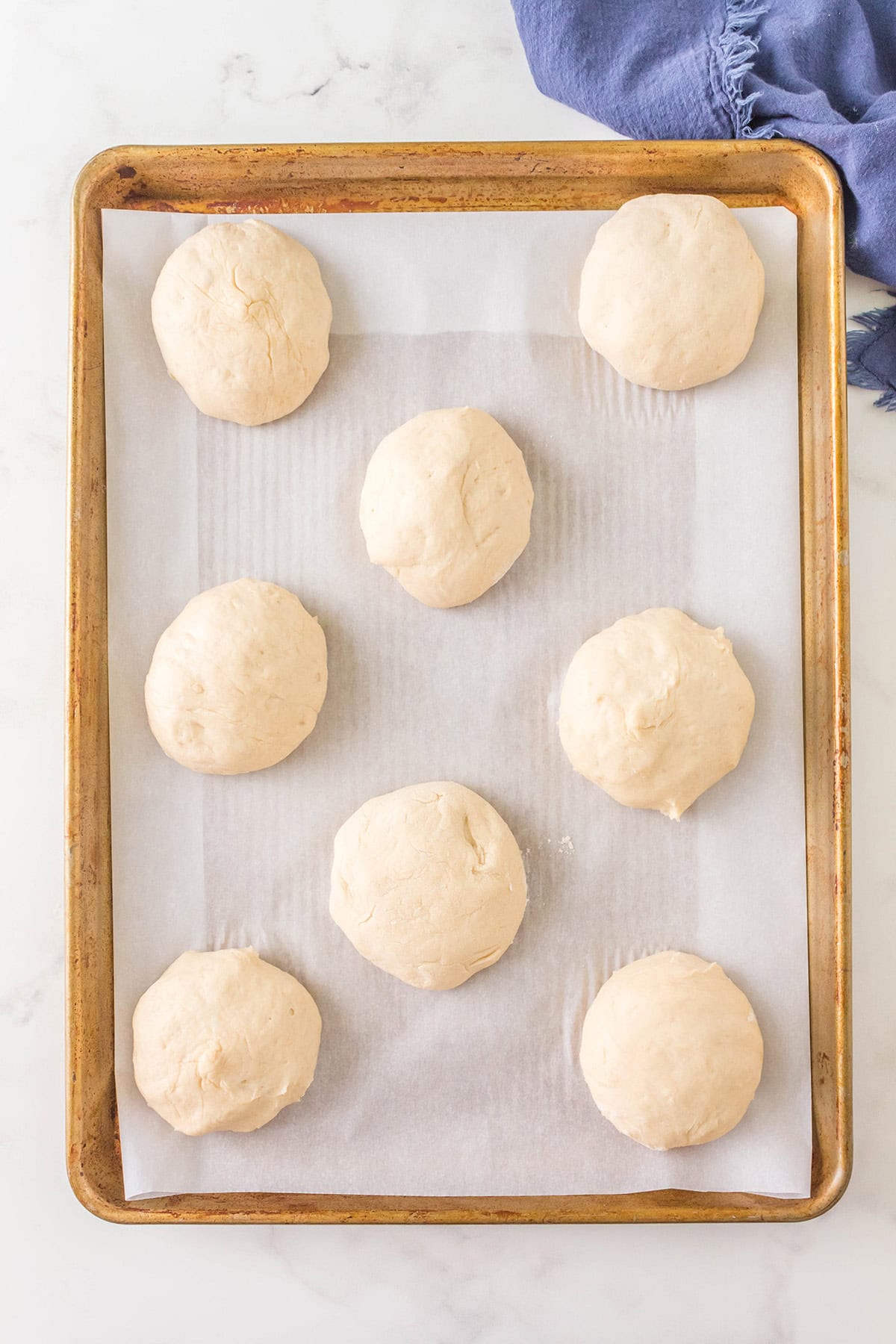 Eight round balls of dough are evenly spaced on a parchment-lined baking sheet. The sheet sits on a light surface with part of a blue cloth visible in the upper right corner.