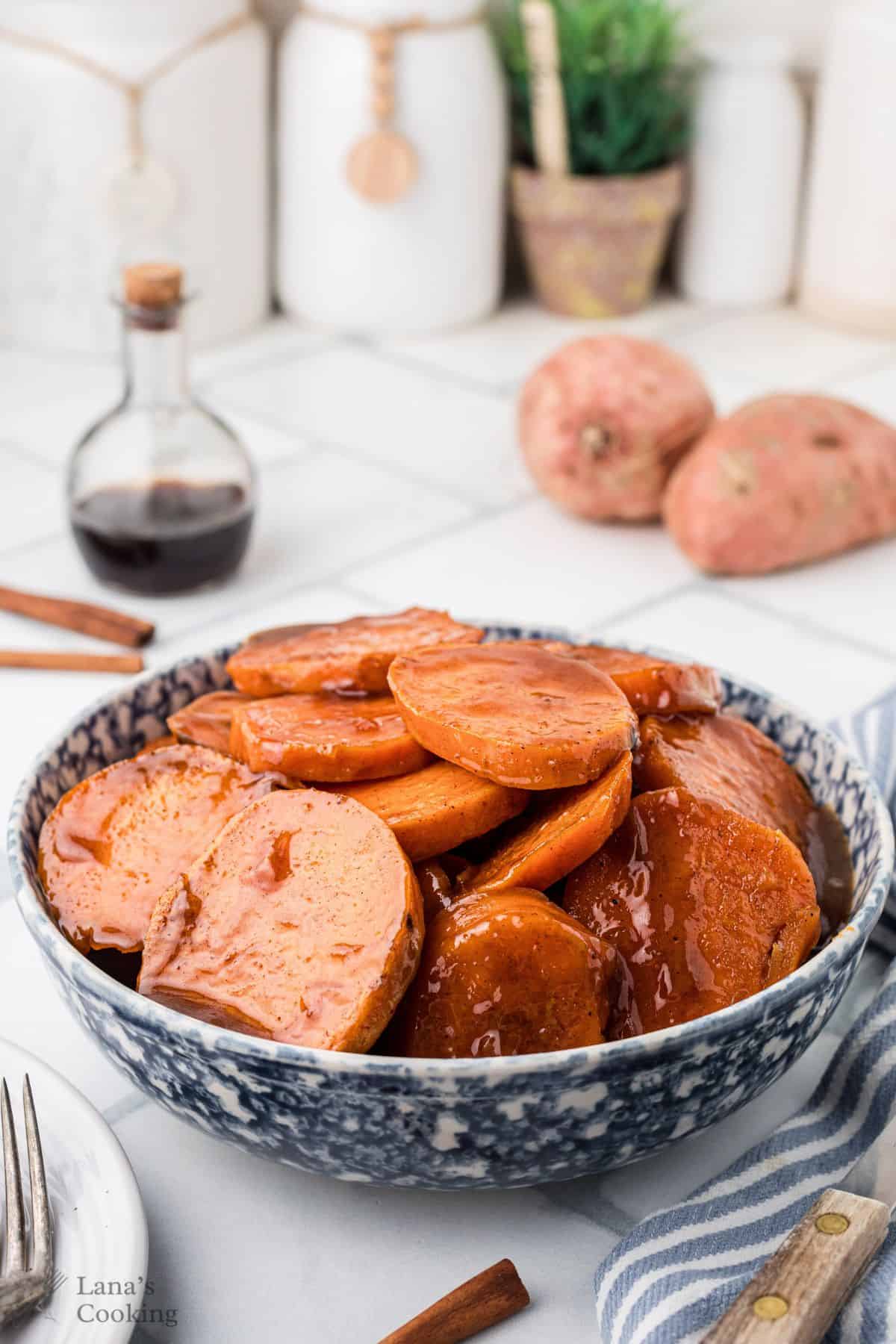 A bowl of glazed candied yams with syrup, sitting on a kitchen counter near fresh sweet potatoes.