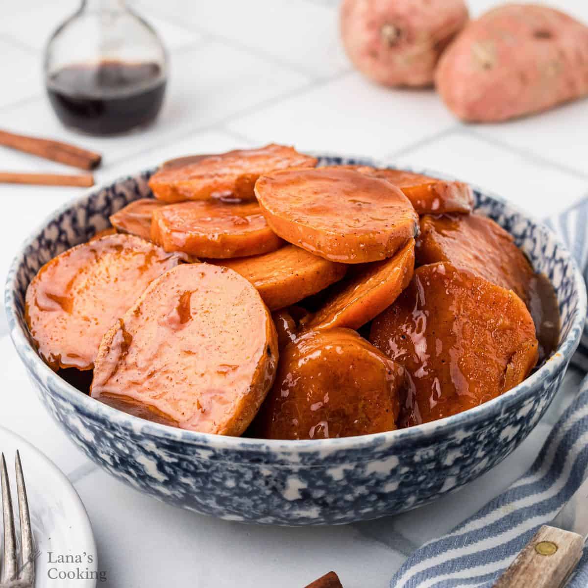 A blue bowl filled with glazed, sliced candied yams on a white table, with sweet potatoes in the background.