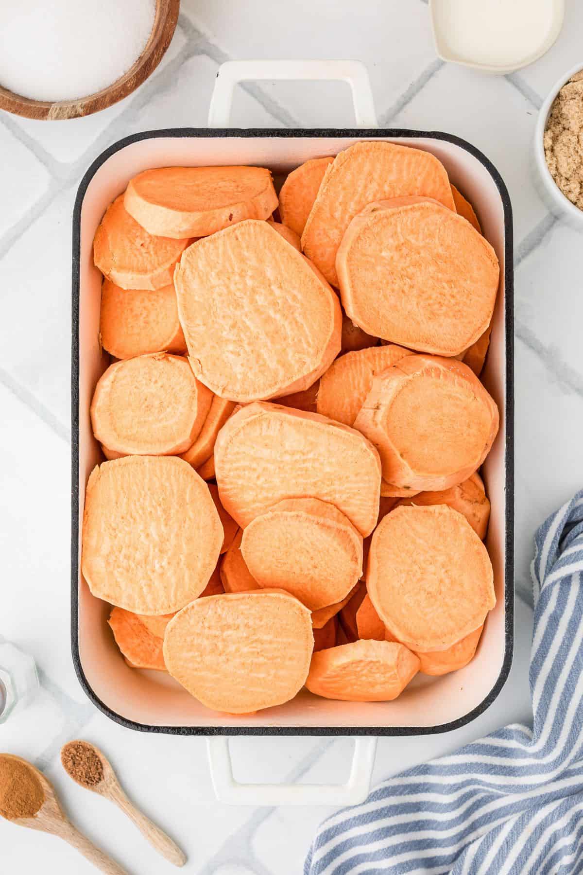 A baking dish filled with raw, sliced sweet potatoes on a white countertop with baking ingredients nearby.