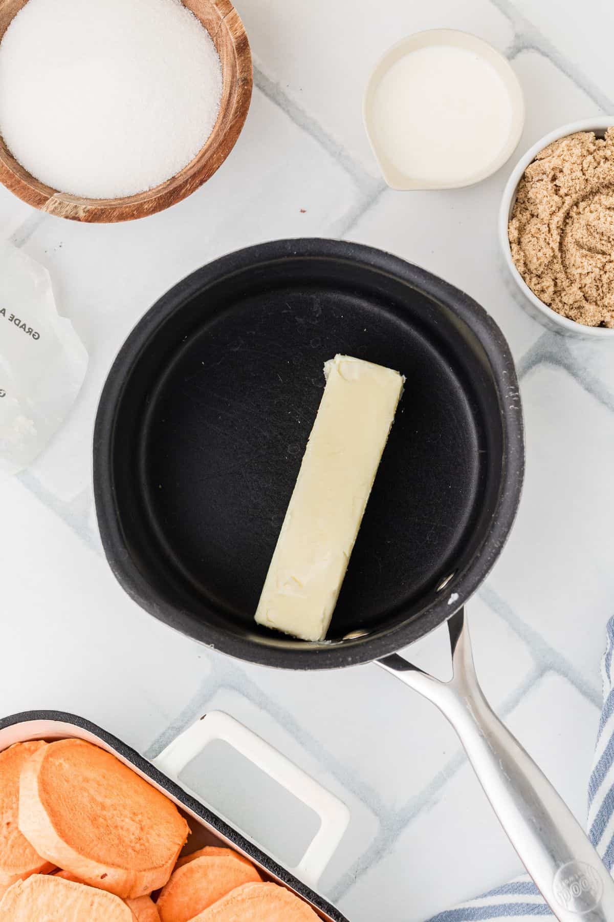 A stick of butter in a saucepan, surrounded by bowls of sugar, brown sugar, cream, and sliced sweet potatoes.
