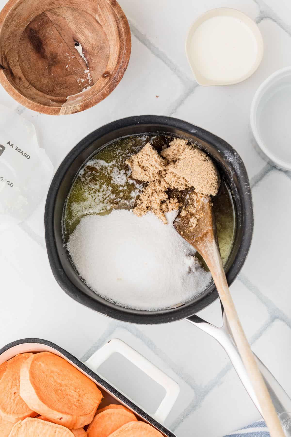 A saucepan with melted butter, white sugar, and brown sugar being mixed with a wooden spoon, next to sweet potatoes.