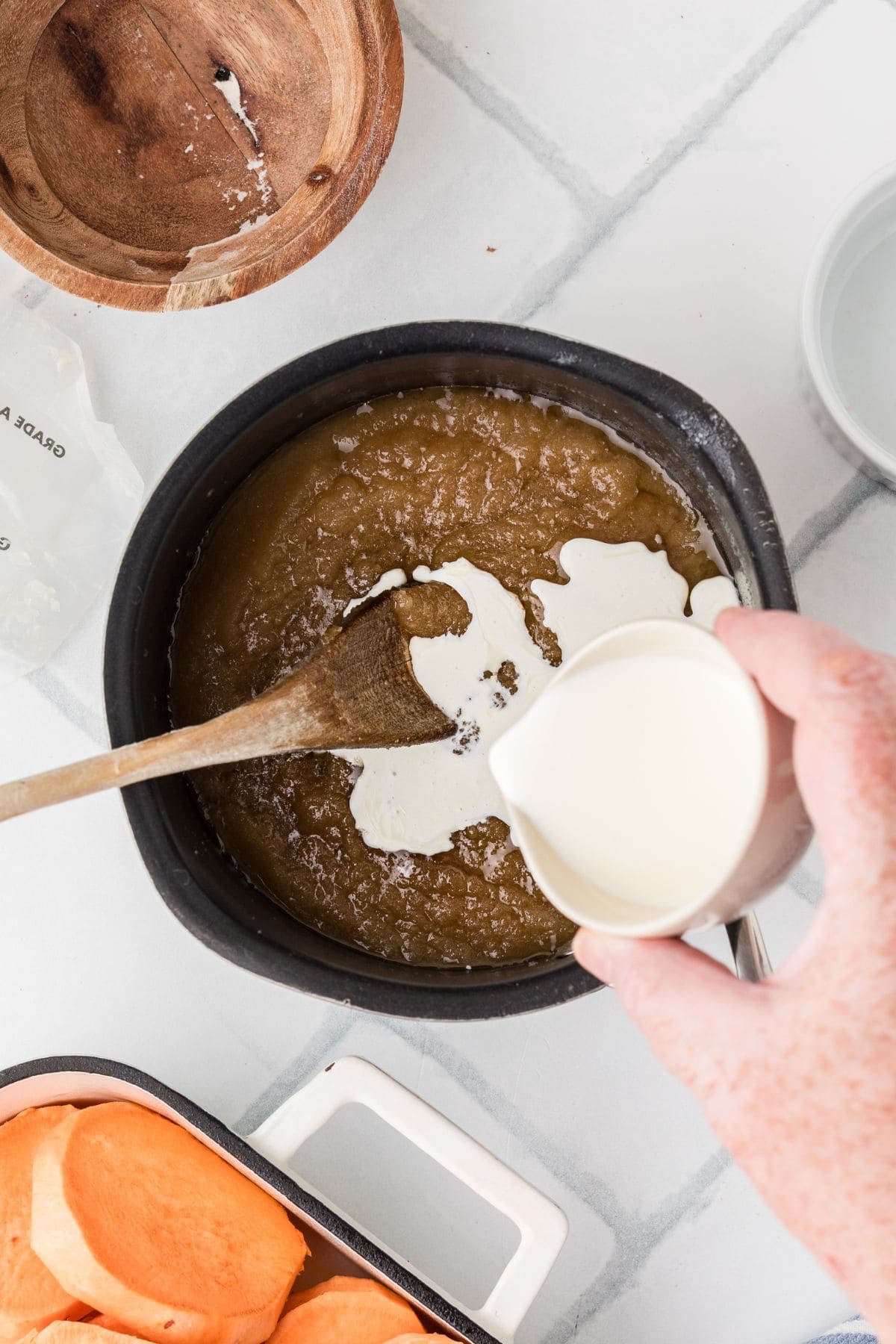A hand pours cream into a saucepan of brown mixture with a wooden spoon, next to sliced sweet potatoes.