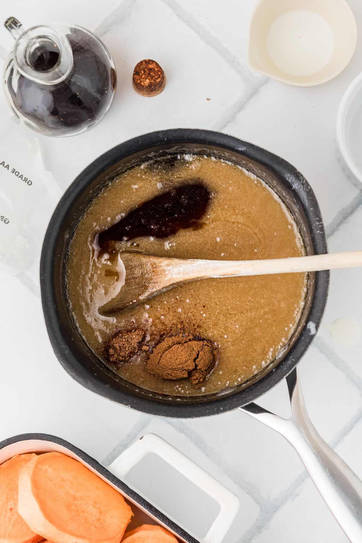 A saucepan with brown sugar and liquid being stirred, surrounded by syrup, sweet potatoes, and bowls on a white surface.
