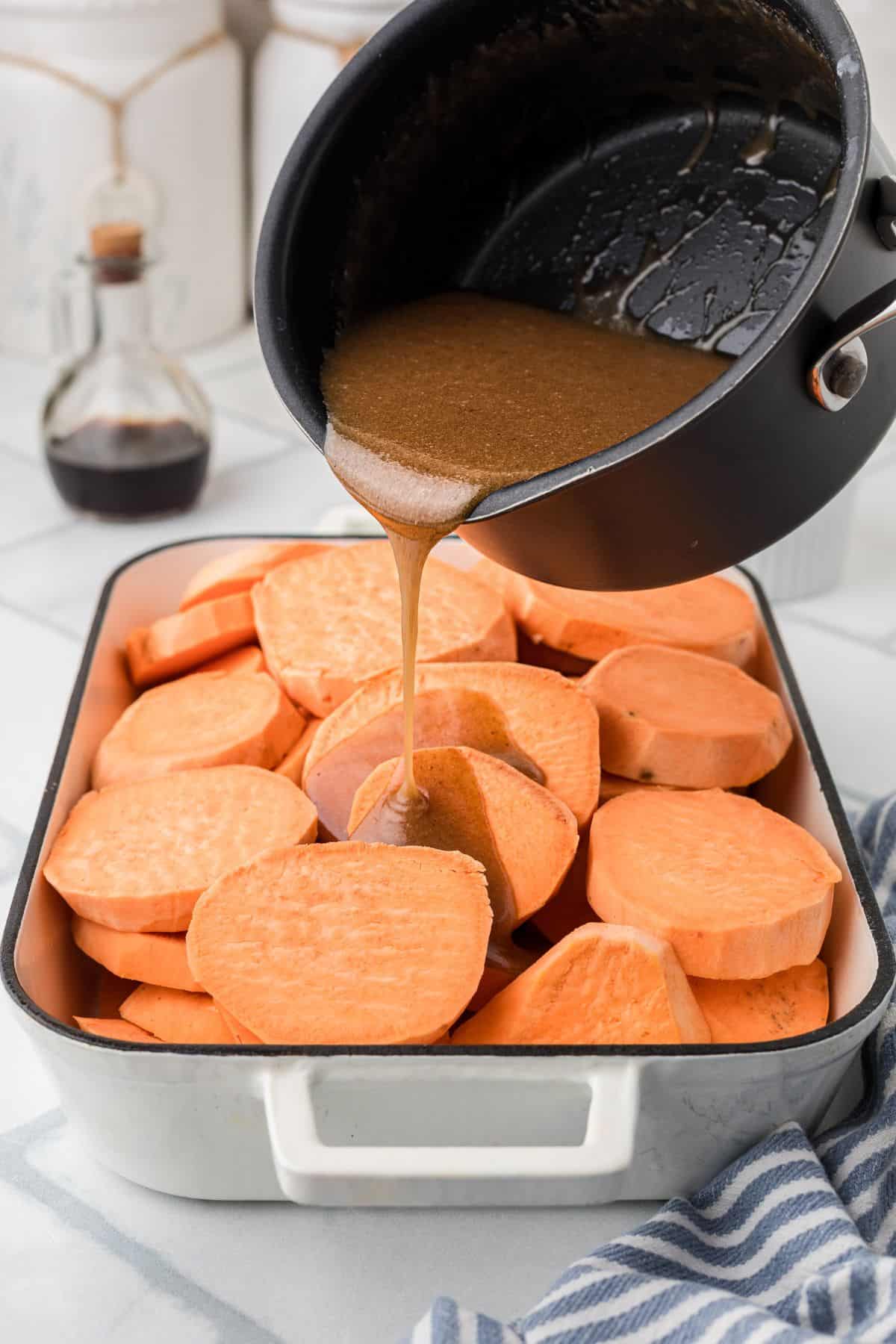 A pot pours brown syrup over sliced sweet potatoes in a white baking dish on a kitchen counter.