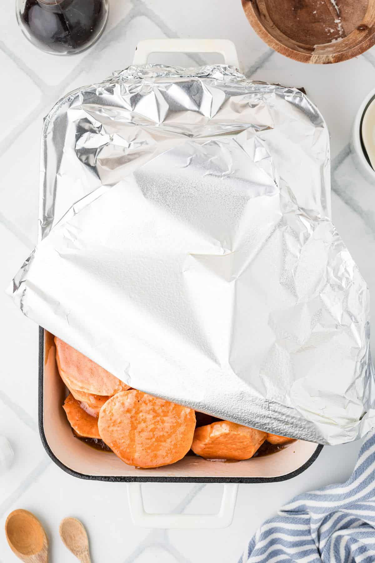A baking dish of sliced sweet potatoes partially covered with aluminum foil on a white countertop.