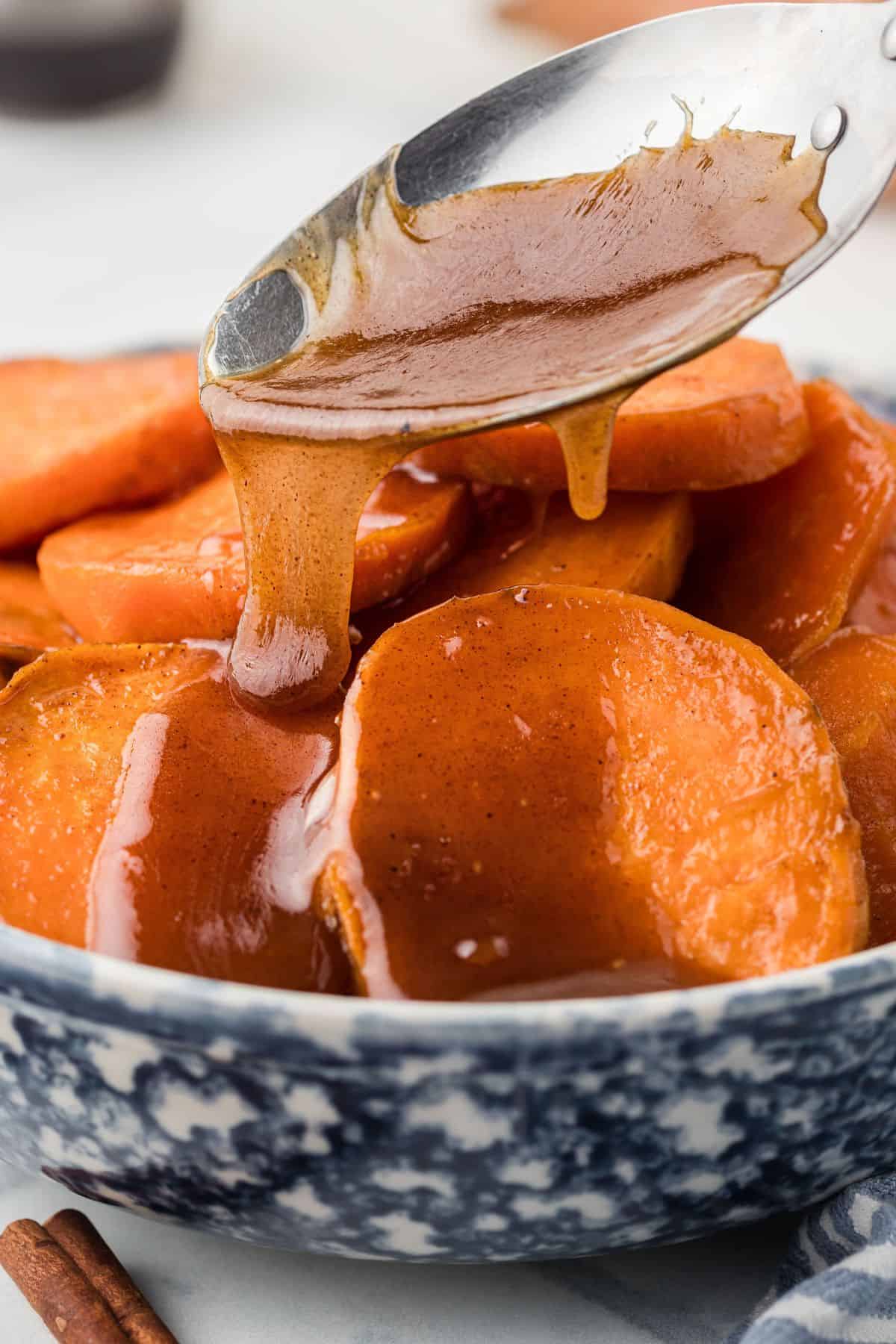 A spoon drizzles syrup over sliced sweet potatoes in a blue and white bowl.