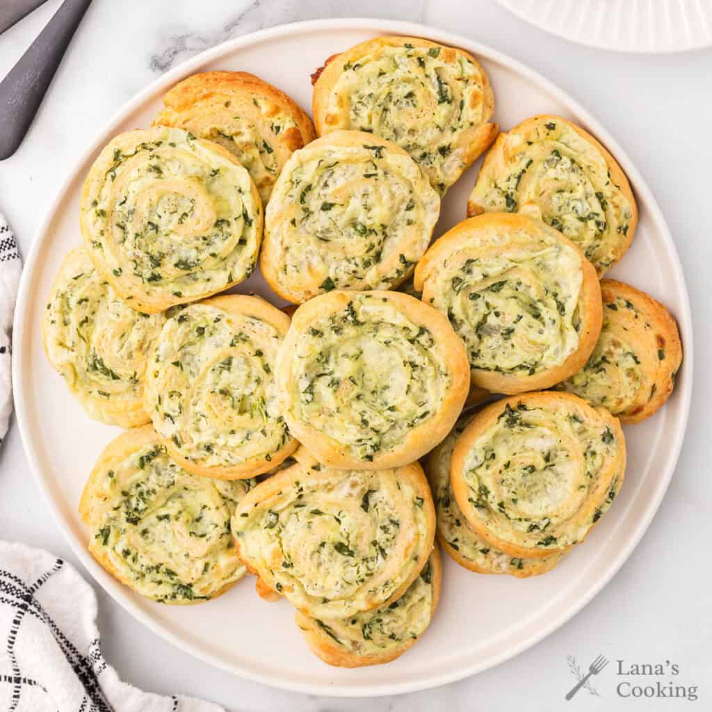 A plate of golden, spiral-shaped spinach and cheese pinwheel pastries arranged neatly on a white plate.