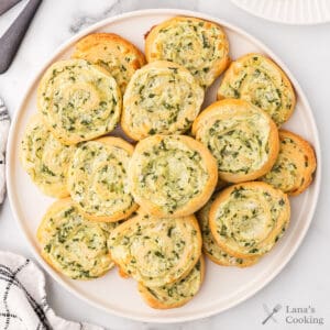 A plate of golden, spiral-shaped spinach and cheese pinwheel pastries arranged neatly on a white plate.